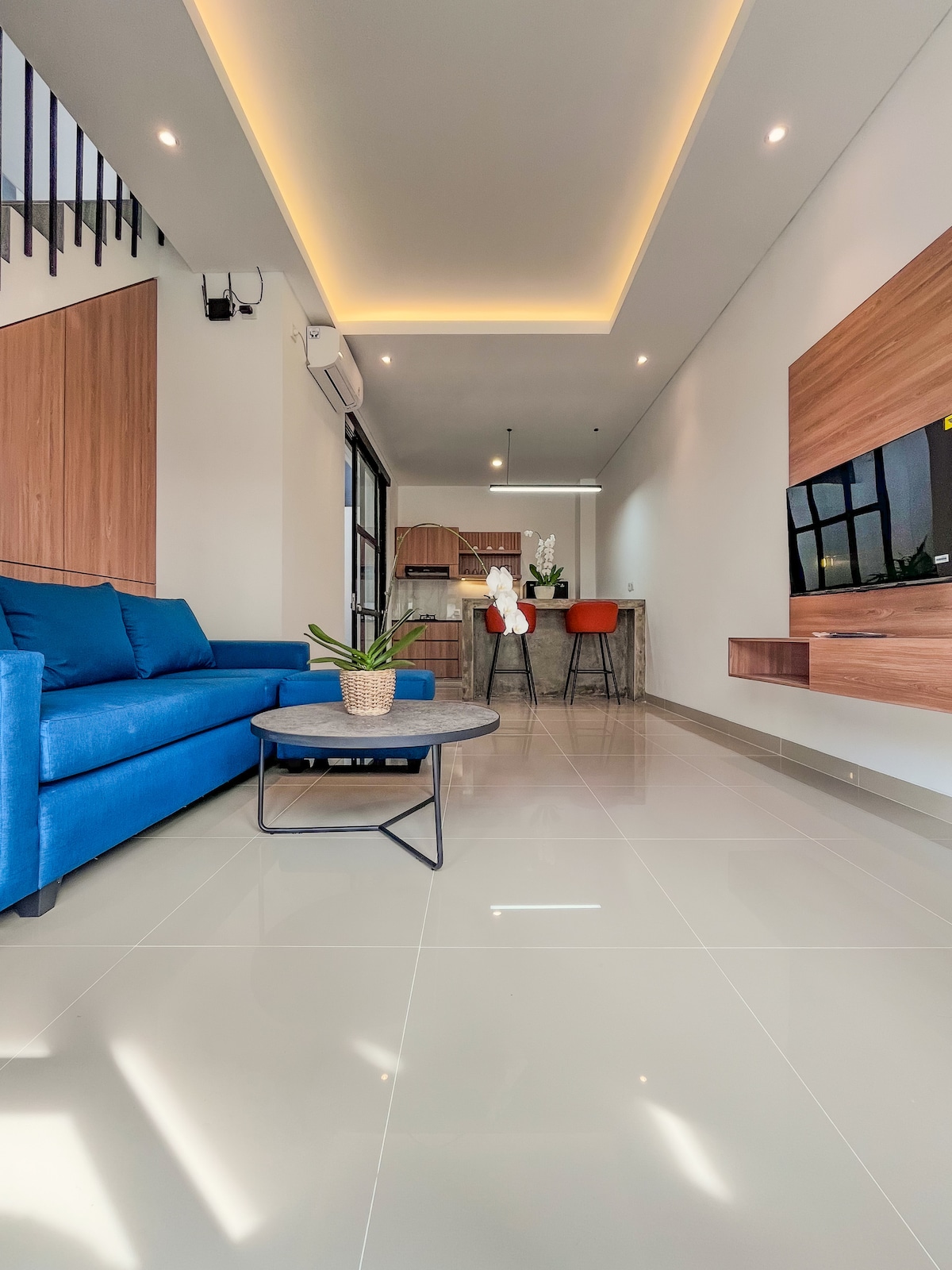 A bright living area features a comfortable blue sofa paired with a round coffee table. Behind, a modern kitchen with bar seating is visible. Light from the window enhances the airy atmosphere of this open-plan space, creating a warm and inviting environment for relaxation.