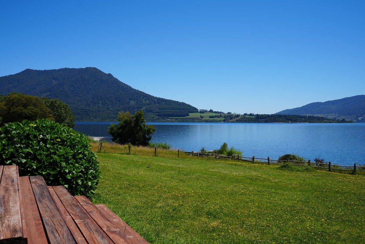 A serene view of a lake surrounded by rolling hills and mountains. Lush greenery lines the water's edge, with a wooden deck visible in the foreground. The clear blue sky reflects on the tranquil lake surface, enhancing the natural beauty of the setting.