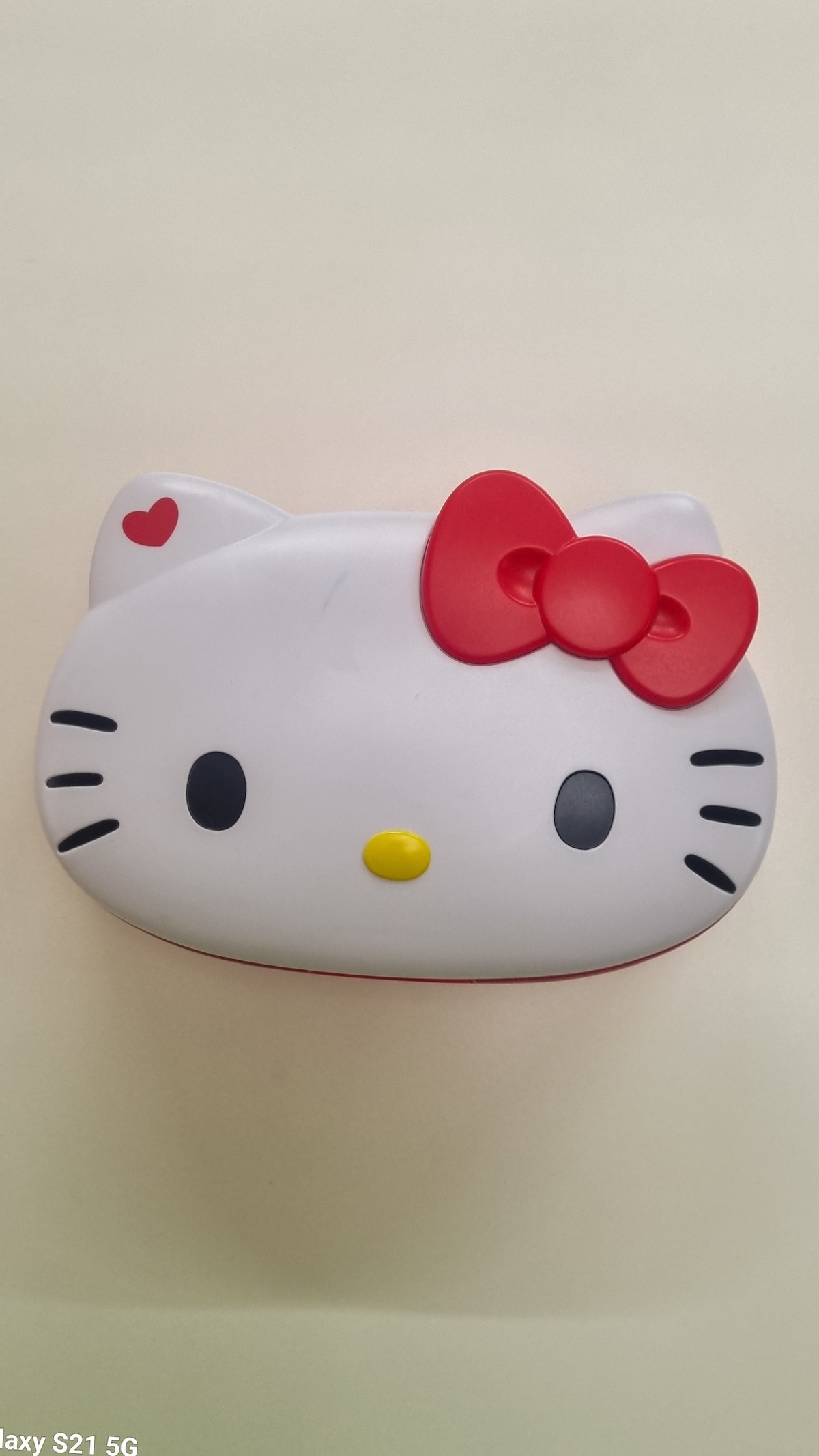 A whimsical Hello Kitty-themed storage container is displayed, featuring a rounded design with prominent facial features. The container is primarily white, adorned with a classic red bow and a small red heart, creating a playful and cheerful appearance.
