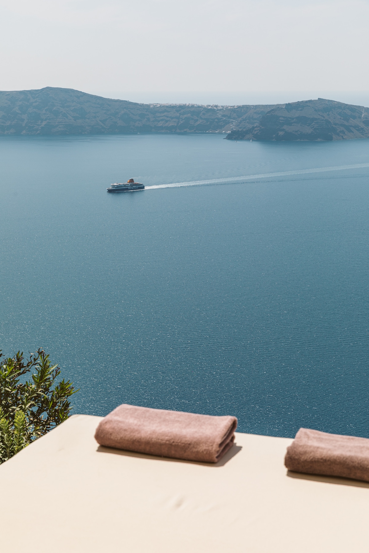 A sunbed is positioned on a terrace, offering a view of a deep blue Aegean sea. Two neatly folded towels are placed on the sunbed, overlooking the water. A distant ferry can be seen traversing the calm sea, framed by rolling green mountains in the background.