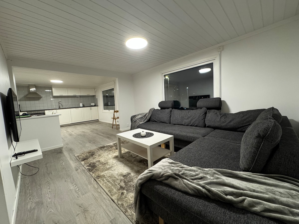 A spacious living area is depicted, featuring a comfortable gray sectional sofa paired with a white coffee table. A soft throw blanket is laid on the sofa. The open layout connects to a modern kitchen visible in the background, flooded with natural light through large windows.