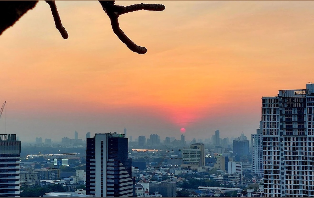 A sunset view showcases a vibrant gradient of orange and pink hues across the sky, with skyscrapers in the foreground and distant city structures outlined against the horizon. The sun is partially visible, creating a serene atmosphere over the urban landscape.