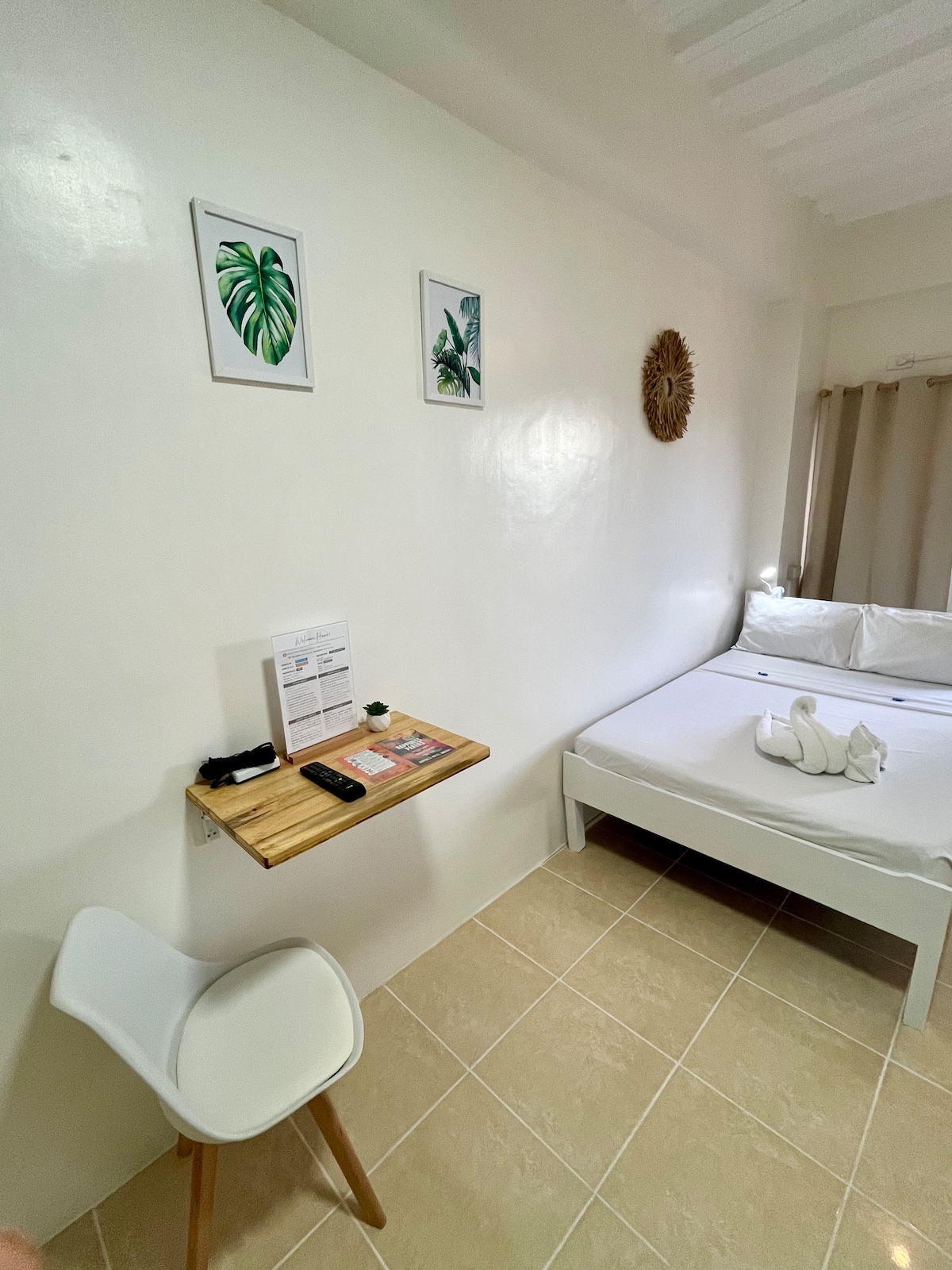 A bright sleeping area features a double-sized bed dressed in white linens, complemented by decorative wall art with leaf motifs. A small wooden table is attached to the wall, providing space for personal items. Natural light filters in through sheer curtains, enhancing the calm ambiance.