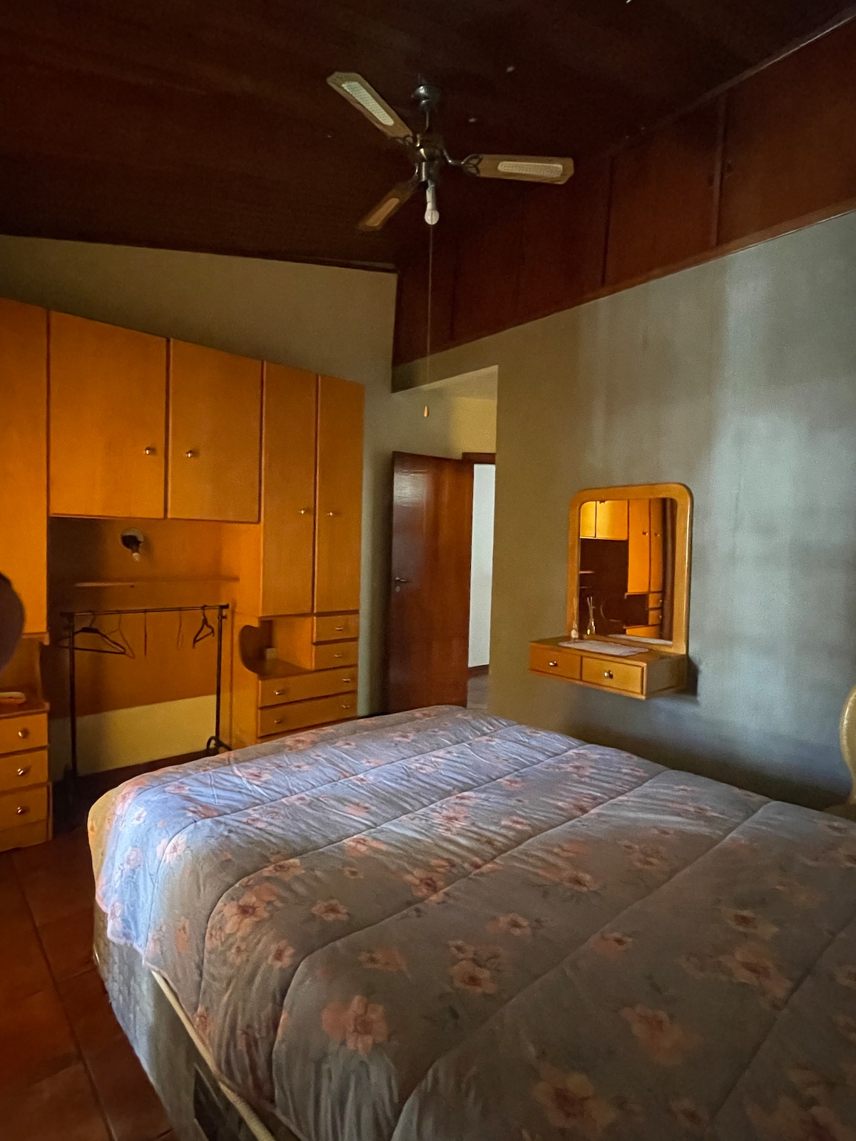 A cozy bedroom is illustrated with a spacious bed covered in floral bedding. Wooden cabinetry lines the walls, providing ample storage. A mirror hangs above a small dresser, reflecting the warm lighting from the exposed ceiling. A ceiling fan is mounted above, contributing to the room's comfort.