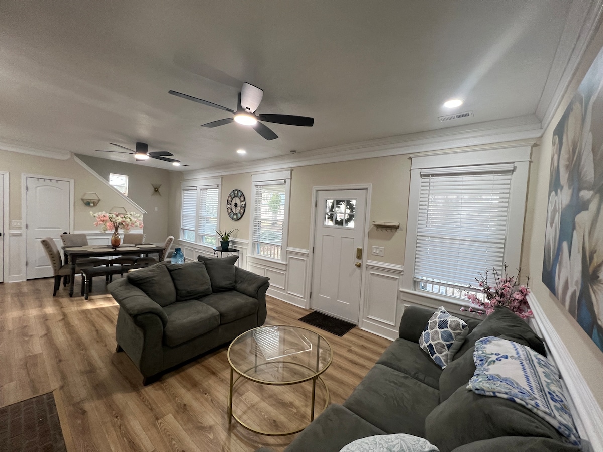 A spacious living area is furnished with a comfortable couch and a glass coffee table. Natural light filters through several windows, illuminating the space. A dining area is visible in the background, featuring a table and chairs. Wall art adds a decorative touch.