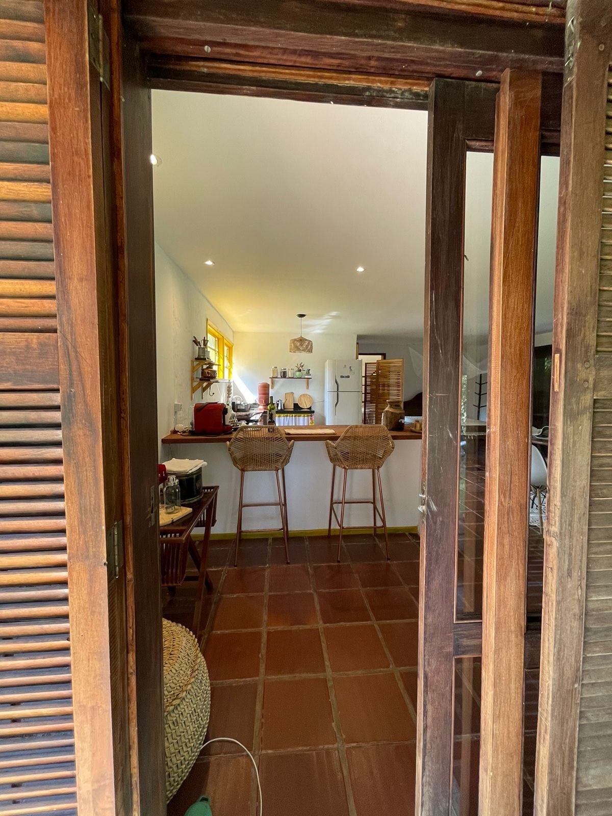 A wooden door stands ajar, revealing a bright kitchen area with a bar equipped with two wicker stools. The space features warm lighting and minimalist decor, complemented by a glimpse of amenities organized on the countertops.