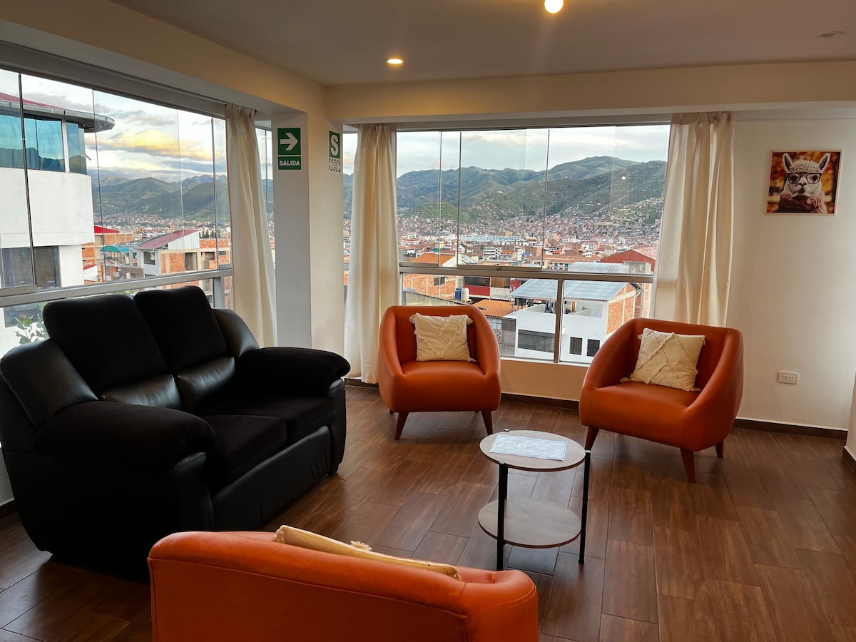 A bright living area offers panoramic views of the mountains and city. Two orange armchairs and a black couch are arranged around a small, round table. Large windows fill the space with natural light, while sheer curtains partially diffuse the brightness.