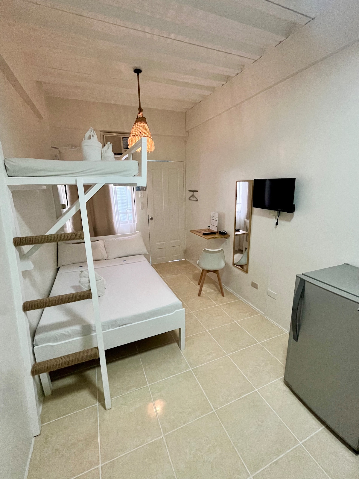 A well-lit room features a double bed positioned beside a bunk bed. A small table with a chair is placed against the wall, while a refrigerator stands nearby. A wall-mounted TV is visible, and a mirror reflects the space, enhancing the room's openness.