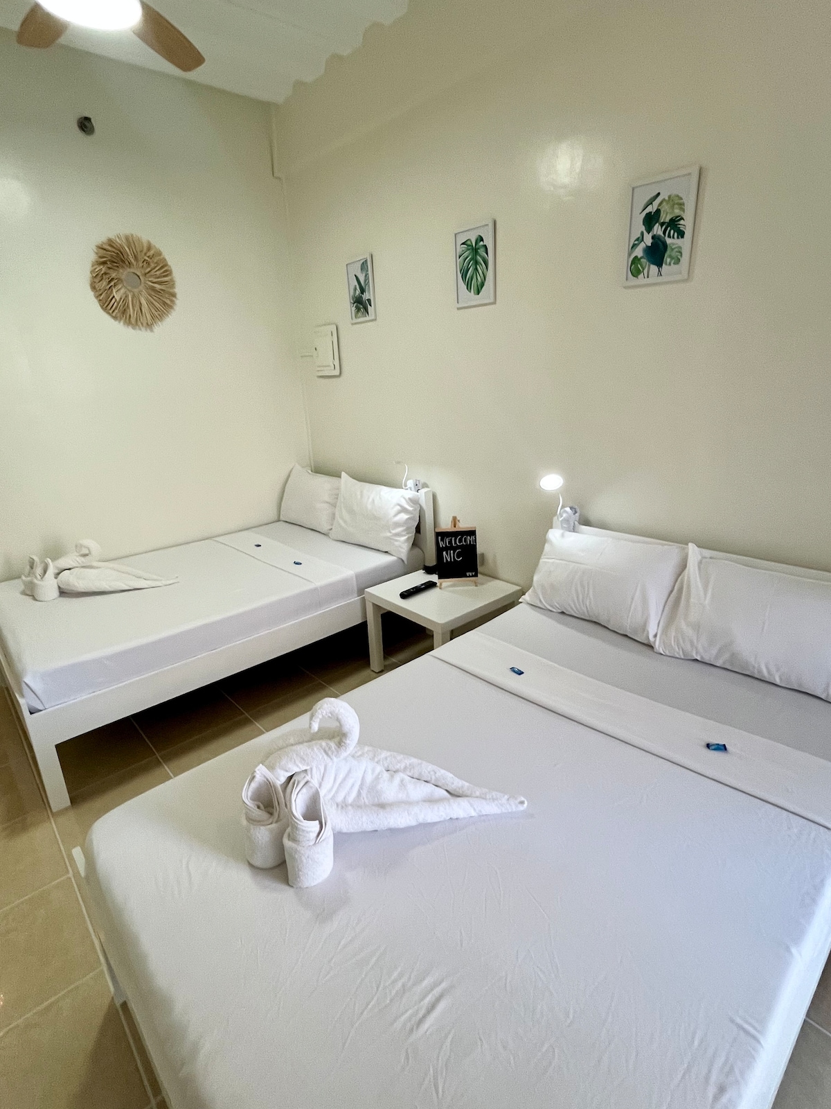 The room features two double beds with white linens, positioned side by side. A nightstand with a lamp is placed between the beds. Minimalist wall art and a decorative piece are displayed above the beds. Natural light is softened by the room's bright walls.