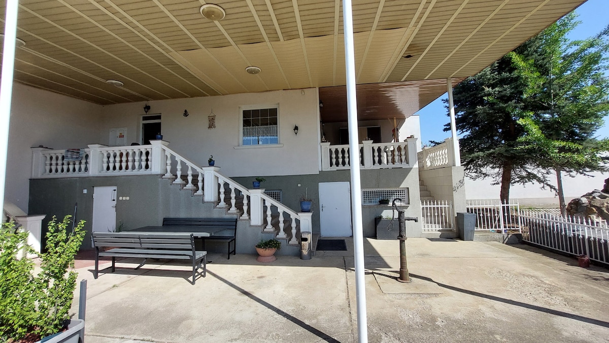 A covered patio area is visible, featuring a stone railing and wide stairs leading to the entrance. Benches are arranged for seating, and planters are positioned along the sides. Trees can be seen in the background, contributing to the outdoor setting.