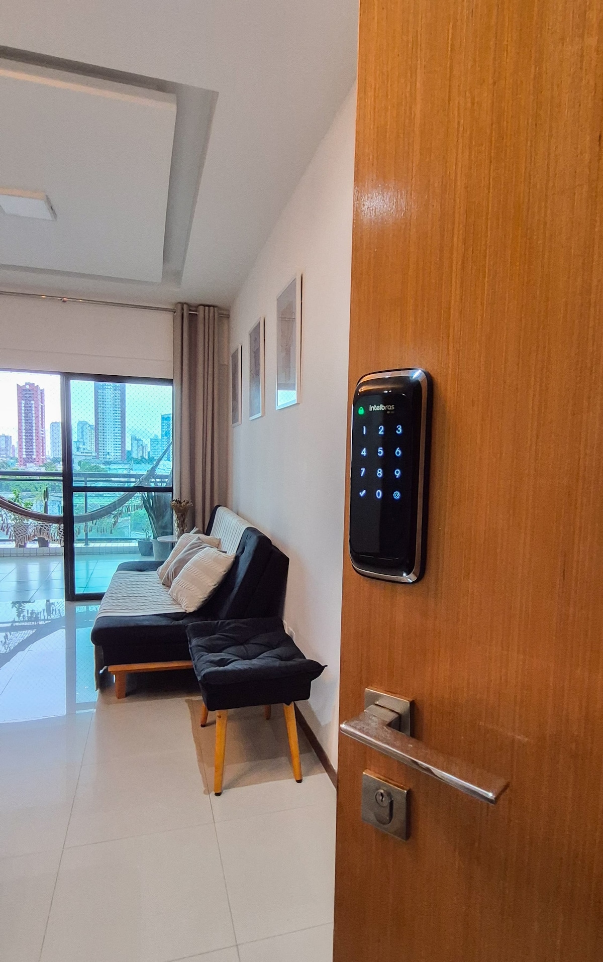 An entry view features a wooden door with a modern electronic lock. A comfortable seating area is visible, with a black sofa and light-colored cushions, welcoming guests into the living space. Natural light flows from the balcony, offering a glimpse of the cityscape beyond.