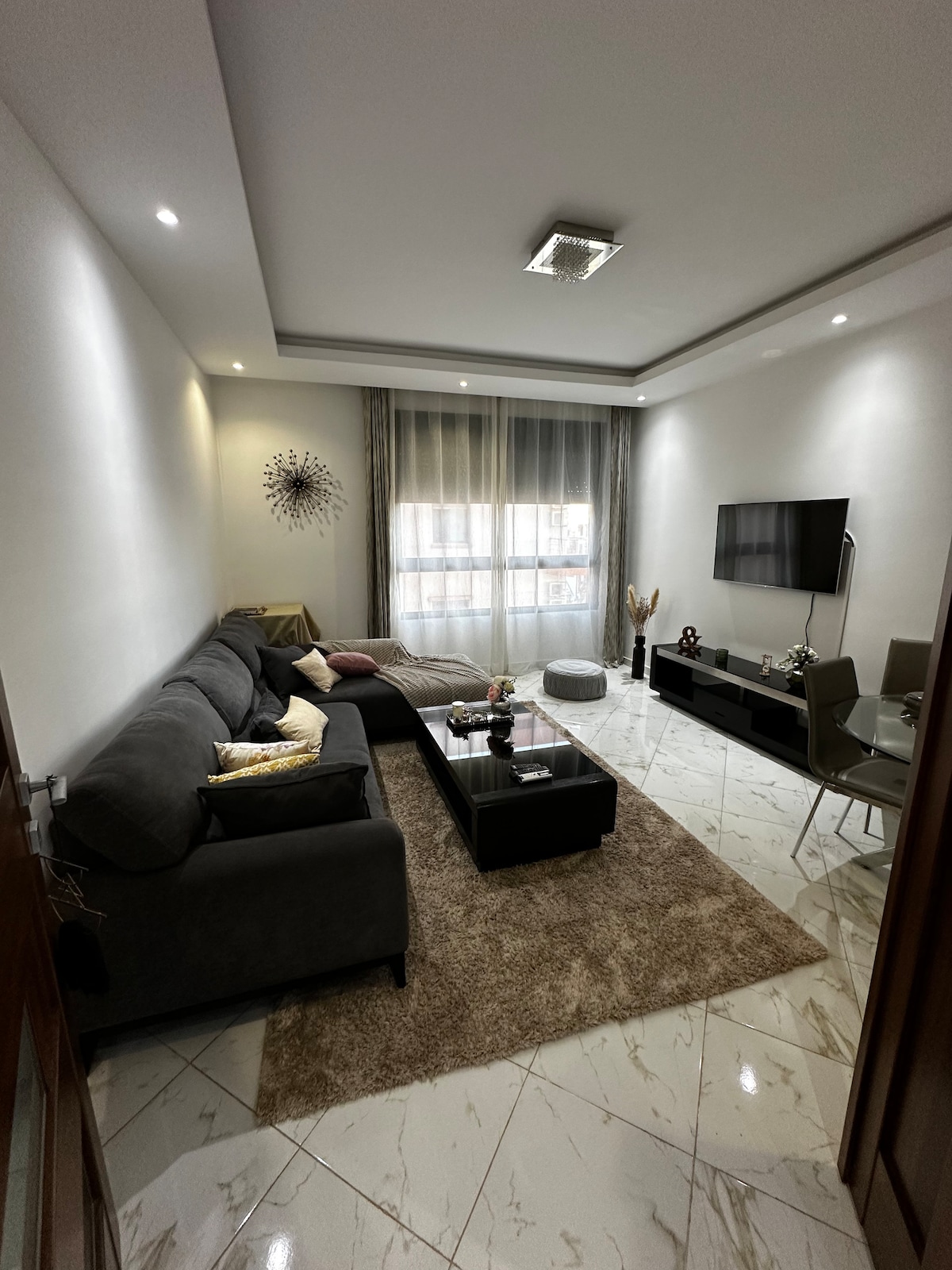 A comfortable living area features a gray sectional sofa adorned with cushions. A glass coffee table sits on a soft area rug, and a flat-screen TV is mounted on the wall. Natural light filters through sheer curtains, illuminating the modern decor.