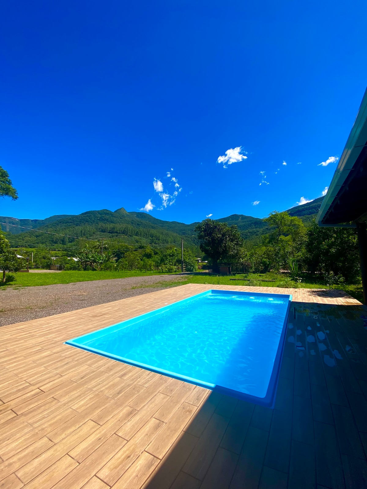 A bright blue swimming pool is set against a backdrop of lush green mountains and a clear blue sky. The pool area features wood-like decking, providing a spacious environment for relaxation and enjoyment.
