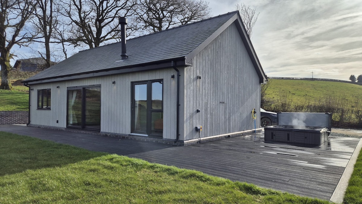 A stylish retreat is captured from the outside, showcasing a modern wooden exterior and large glass doors. A private hot tub is situated on the spacious decking area, surrounded by well-maintained grass and framed by the serene countryside landscape.
