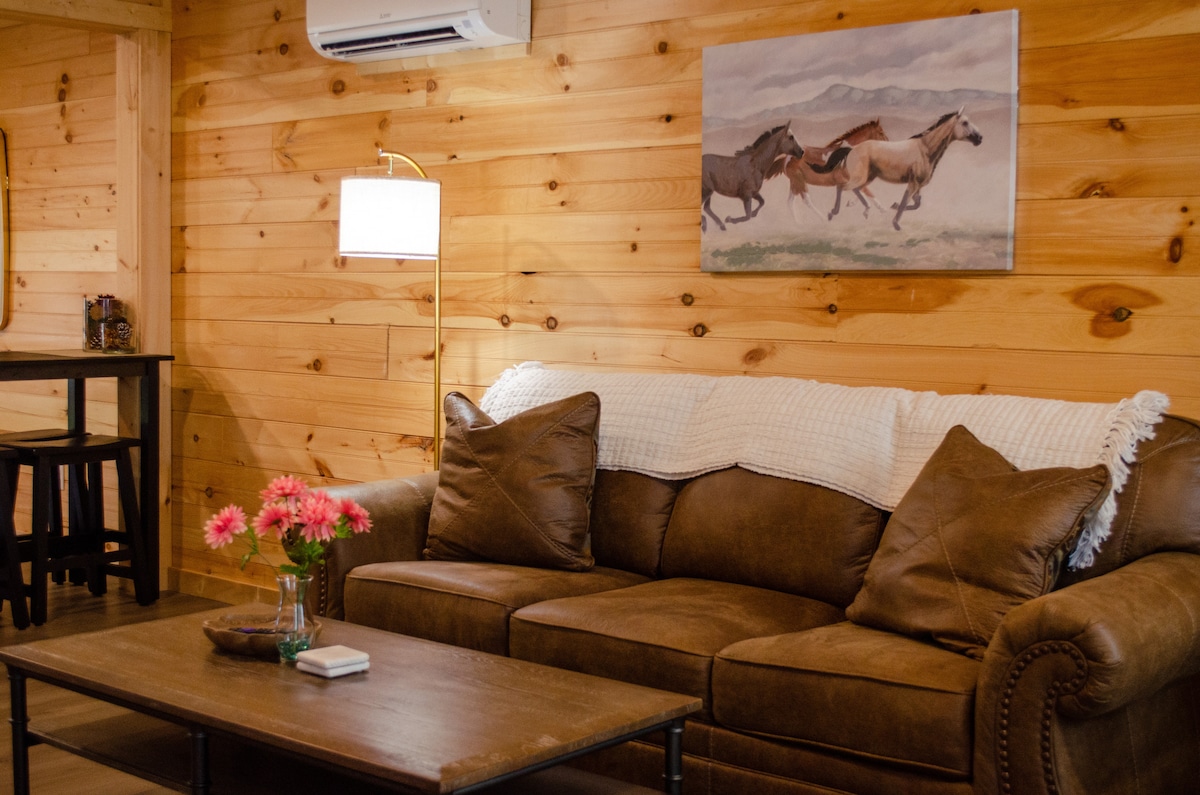 A warm living area features a brown leather couch adorned with plush cushions. A coffee table sits in front, accompanied by a vase of pink flowers. A painting of galloping horses decorates the wooden wall, while soft lighting is provided by a contemporary lamp.
