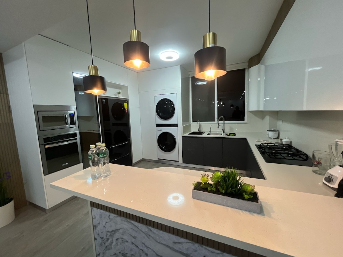 A modern kitchen features sleek cabinetry and black accents. Three pendant lights illuminate the space above a light countertop. Appliances include a built-in oven, a stove, and a dishwasher. A small arrangement of greenery adds a touch of nature to the contemporary design.