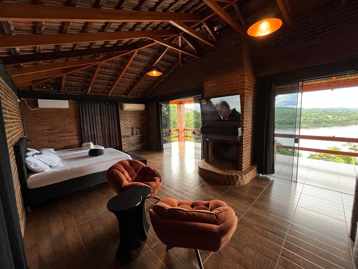 A spacious sleeping area is designed with rustic wooden beams and warm lighting. A large window offers a view of the surrounding landscape. A smart TV is mounted on the wall, and two armchairs are positioned near a cozy fireplace.