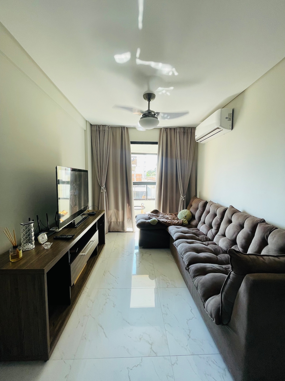 A light-filled living area features a plush brown sectional sofa and a wooden TV stand. The space is adorned with soft curtains that frame a window, allowing natural light to enhance the room. A ceiling fan provides additional comfort.