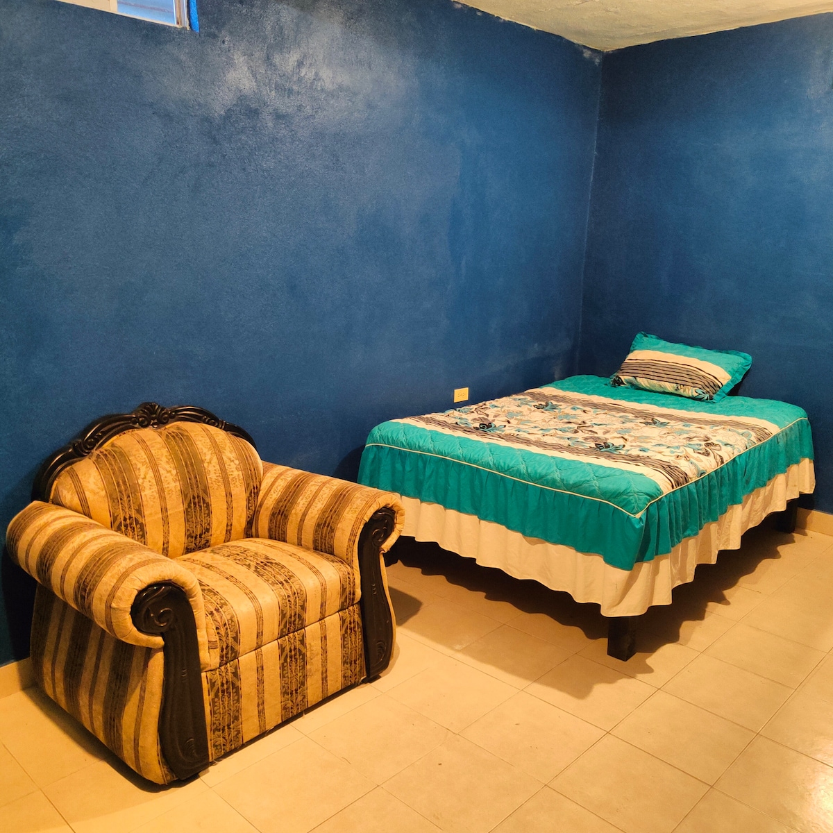 A cozy room is featured with vibrant blue walls. A spacious bed is dressed with a colorful quilt, while a striped armchair adds a touch of comfort. Neutral tile flooring enhances the bright ambiance of the space.