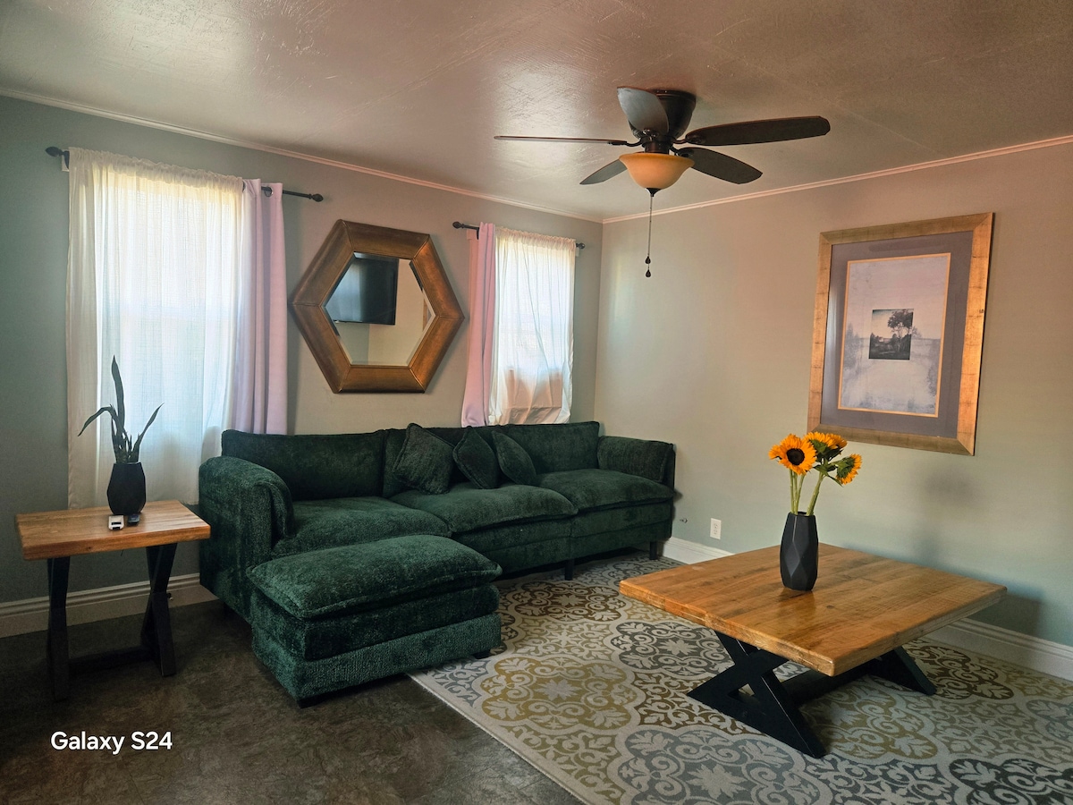 A well-furnished living area features a green sectional sofa, paired with a wooden coffee table and a small side table. Bright sunflowers are placed on the side table, and light filters through sheer curtains. A decorative mirror and framed wall art add visual interest.
