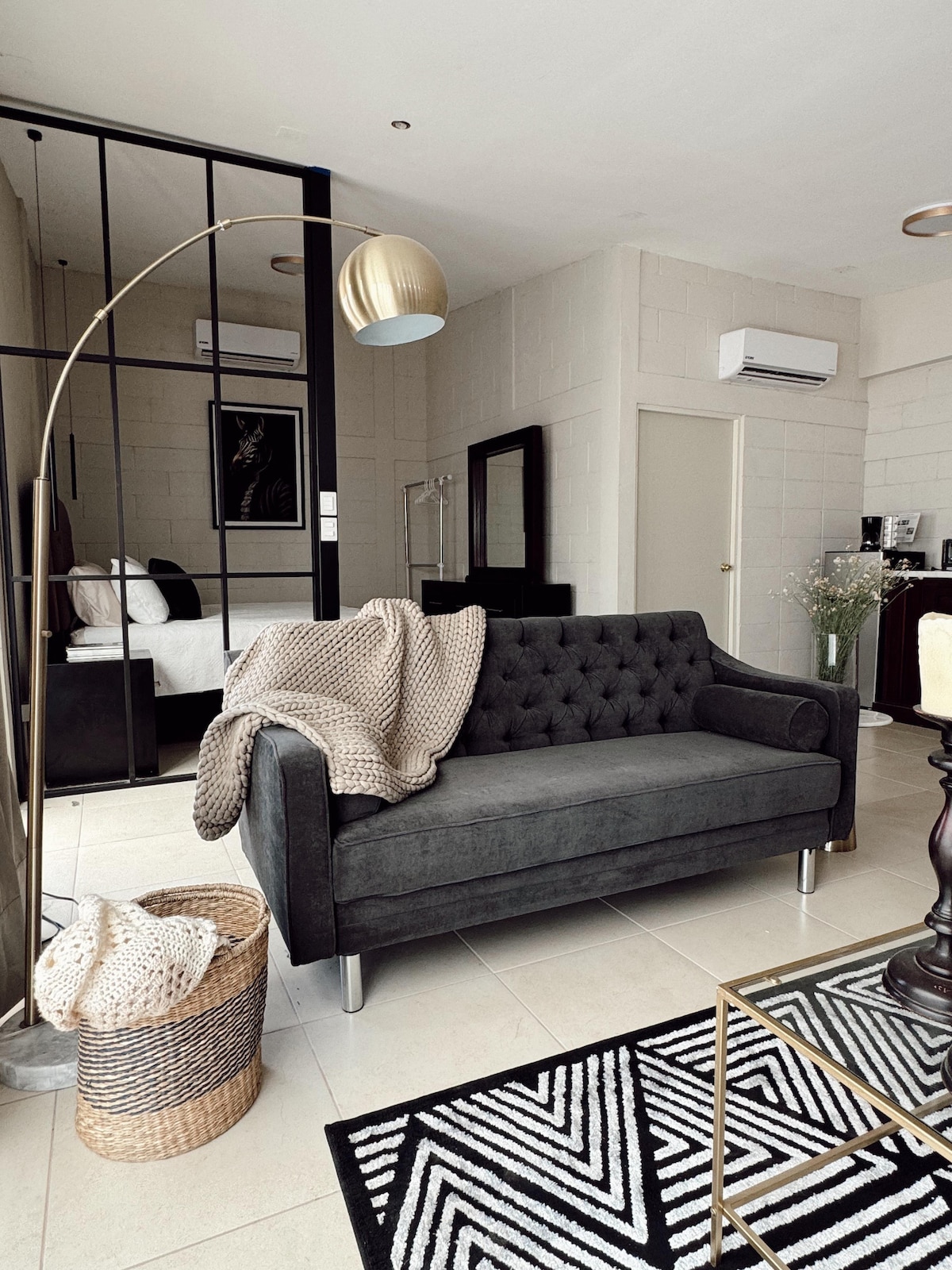A modern living area features a dark tufted sofa adorned with a beige knitted throw. A standing lamp provides soft lighting nearby, while a woven basket adds texture. The background reveals a glimpse of a bedroom with a large bed and minimalist decor.