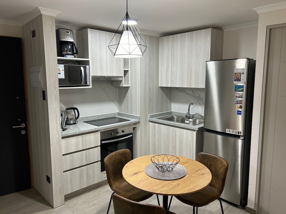 A modern kitchen area is equipped with stainless steel appliances and wooden cabinetry. A round dining table with four chairs is positioned in the center, complemented by a geometric pendant light above. The countertop features a bowl and various kitchen essentials.