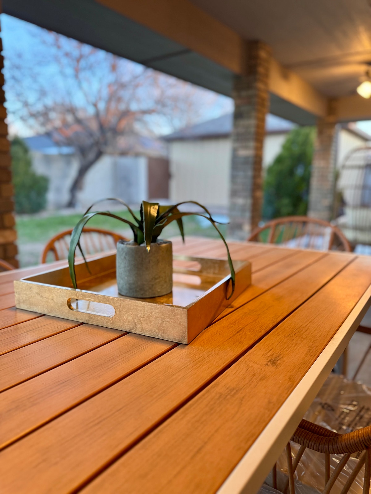 A wooden dining table is featured with a minimalist tray holding a small potted plant. Warm lighting enhances the outdoor area, which includes a backdrop of greenery and a hint of distant structures under a calming sky.