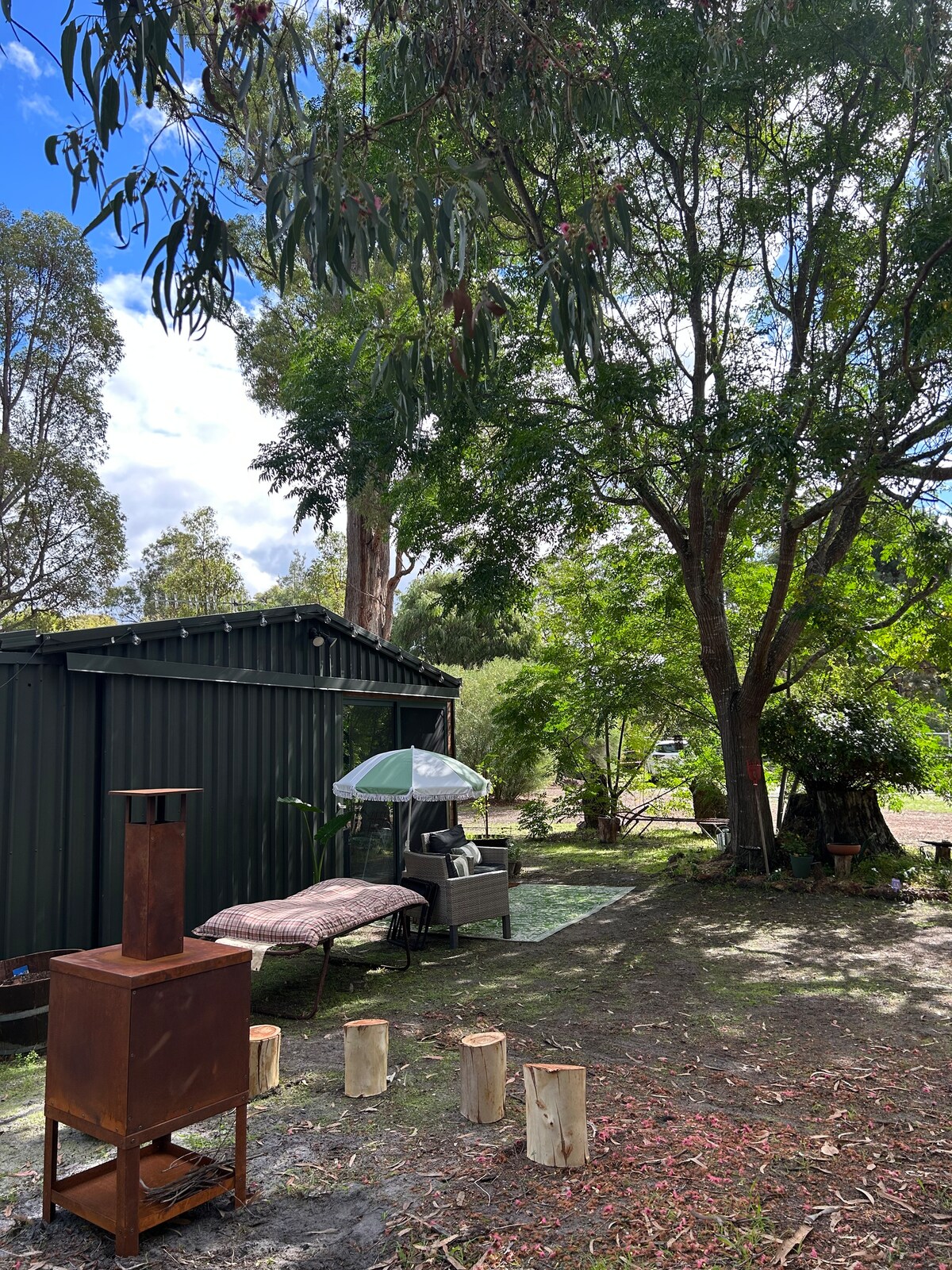 An outdoor area is shown, featuring a seating arrangement under a shaded umbrella next to a studio. A rust-colored fire pit stands in the foreground, surrounded by rustic wooden logs. Lush greenery and trees create a serene backdrop, enhancing the natural setting.
