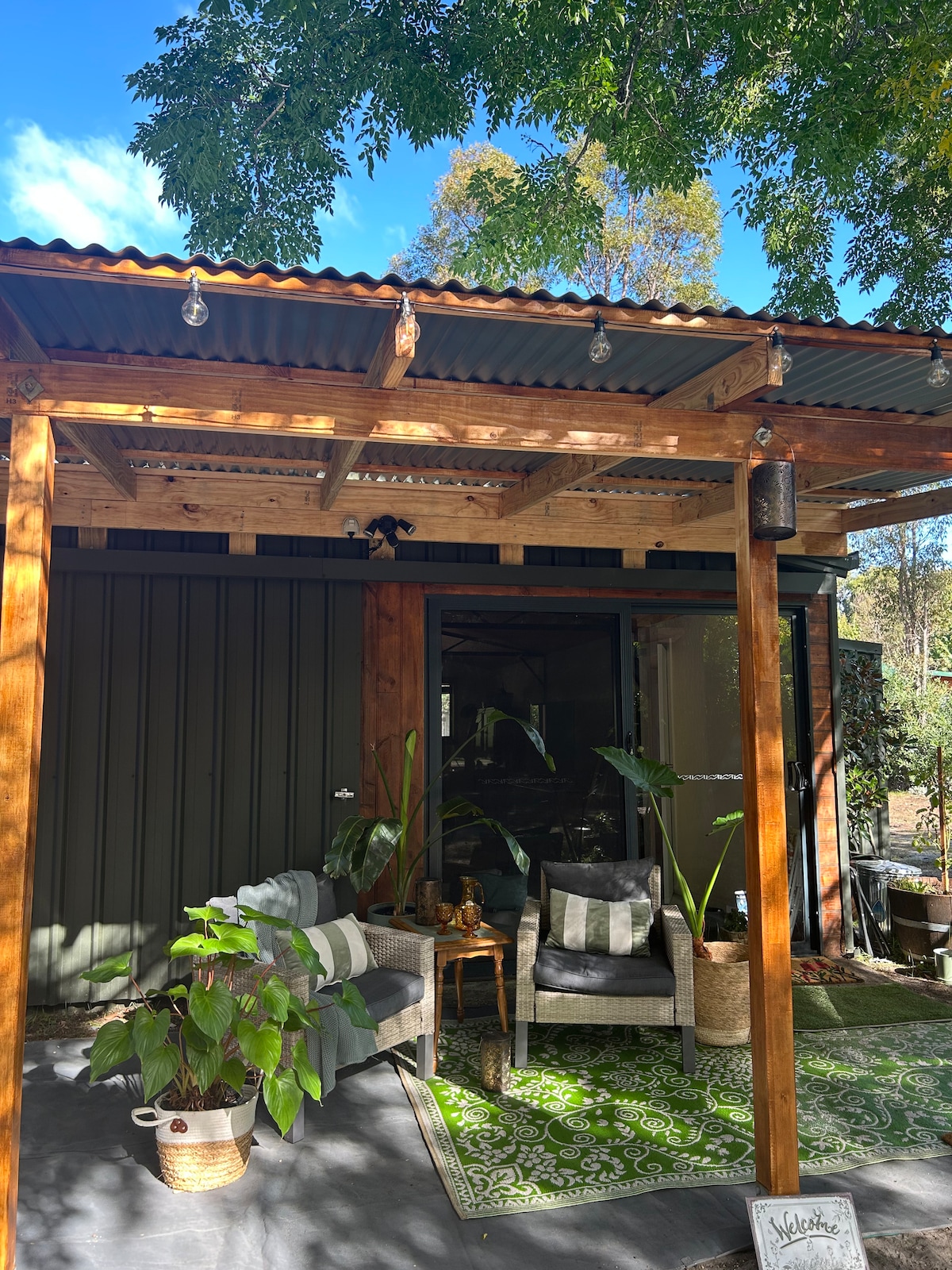 An inviting outdoor seating area is framed by a wooden pergola. Two comfortable chairs with cushions sit on a green patterned rug, surrounded by potted plants. The space is bathed in natural light, with decorative string lights overhead providing a cozy ambiance.