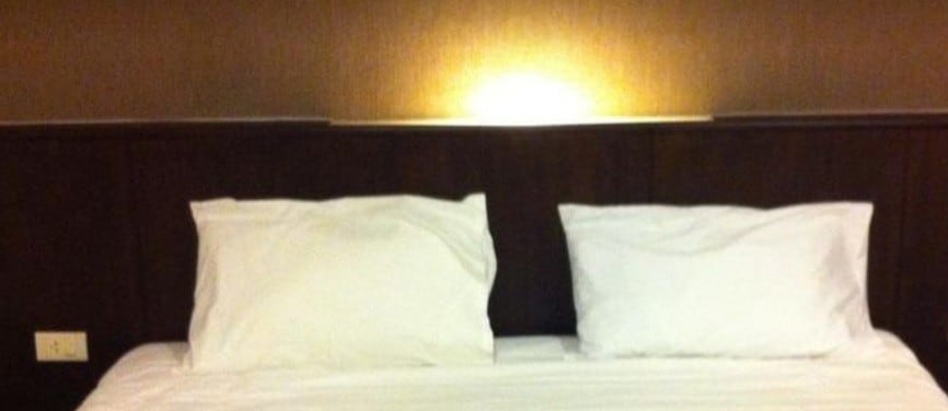 A spacious bed is centered against a warm, textured wall. Two white pillows are positioned neatly at the head of the bed, illuminated by soft lighting above, creating a calming ambiance.