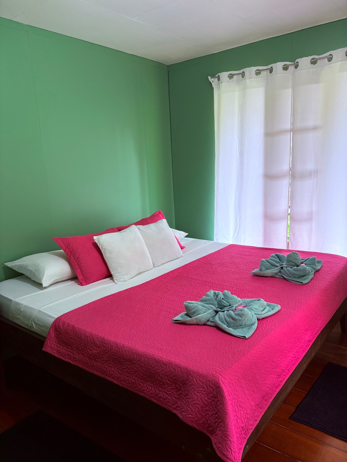 A sleeping area features a spacious bed topped with a vibrant pink coverlet. Coordinating teal towels are artfully arranged on the bed. Natural light enters through a nearby window, partially covered with sheer curtains, enhancing the room's bright green walls.