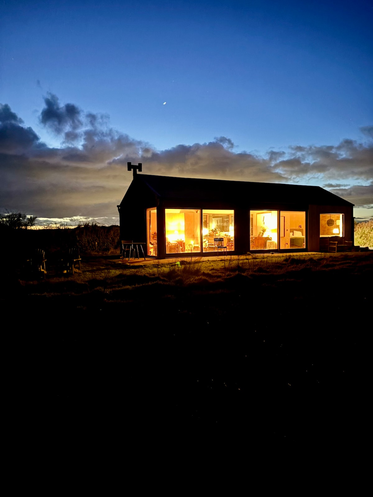A contemporary studio retreat is illuminated at dusk, showcasing large glass doors that provide a glimpse of the inviting interior. The structure is surrounded by natural landscape, with a soft twilight sky and clouds, enhancing the serene setting.