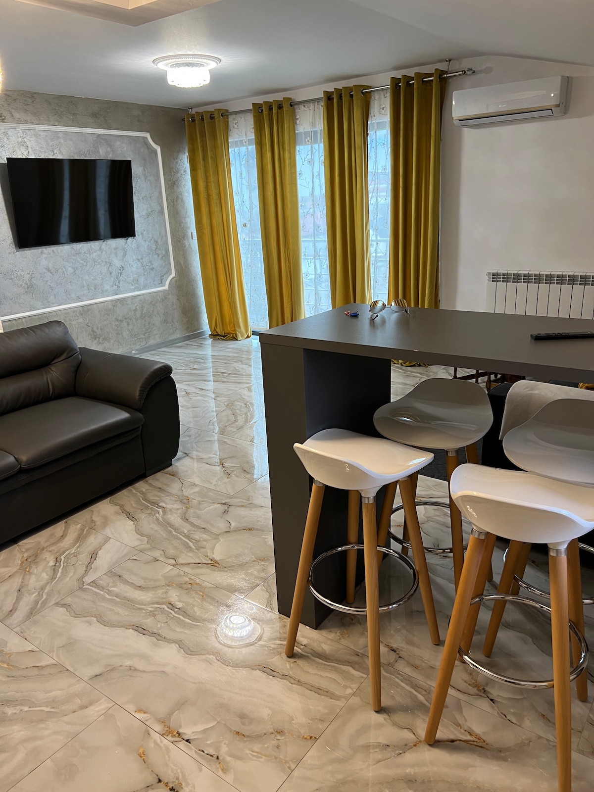 A modern living space features sleek, glossy tile flooring and a comfortable leather sofa. A small dining area with tall, white bar stools is positioned next to a dark countertop. Large windows are dressed with vibrant yellow curtains, allowing natural light to fill the room.
