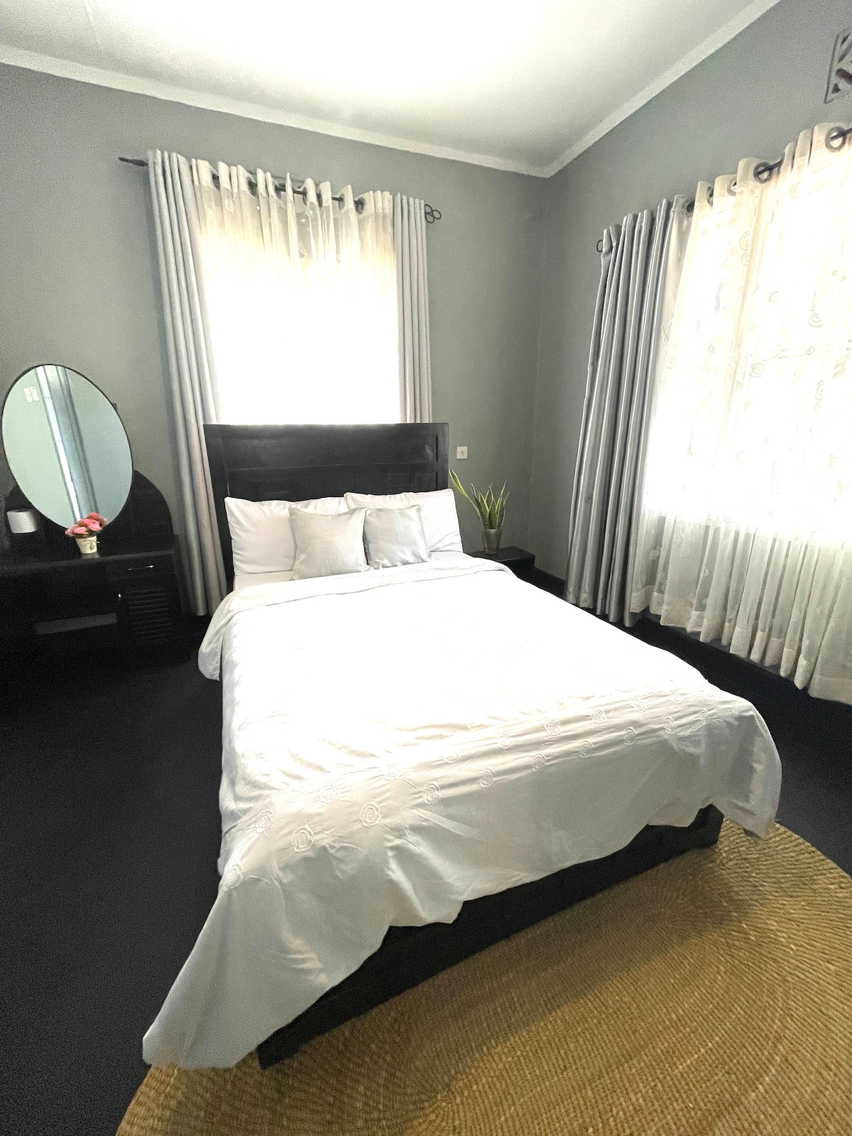 A serene bedroom showcases a spacious bed covered with white linens. A mirror is positioned near the bed, reflecting the room's light. Light-colored curtains frame the window, and a natural fiber rug adds warmth to the dark flooring.