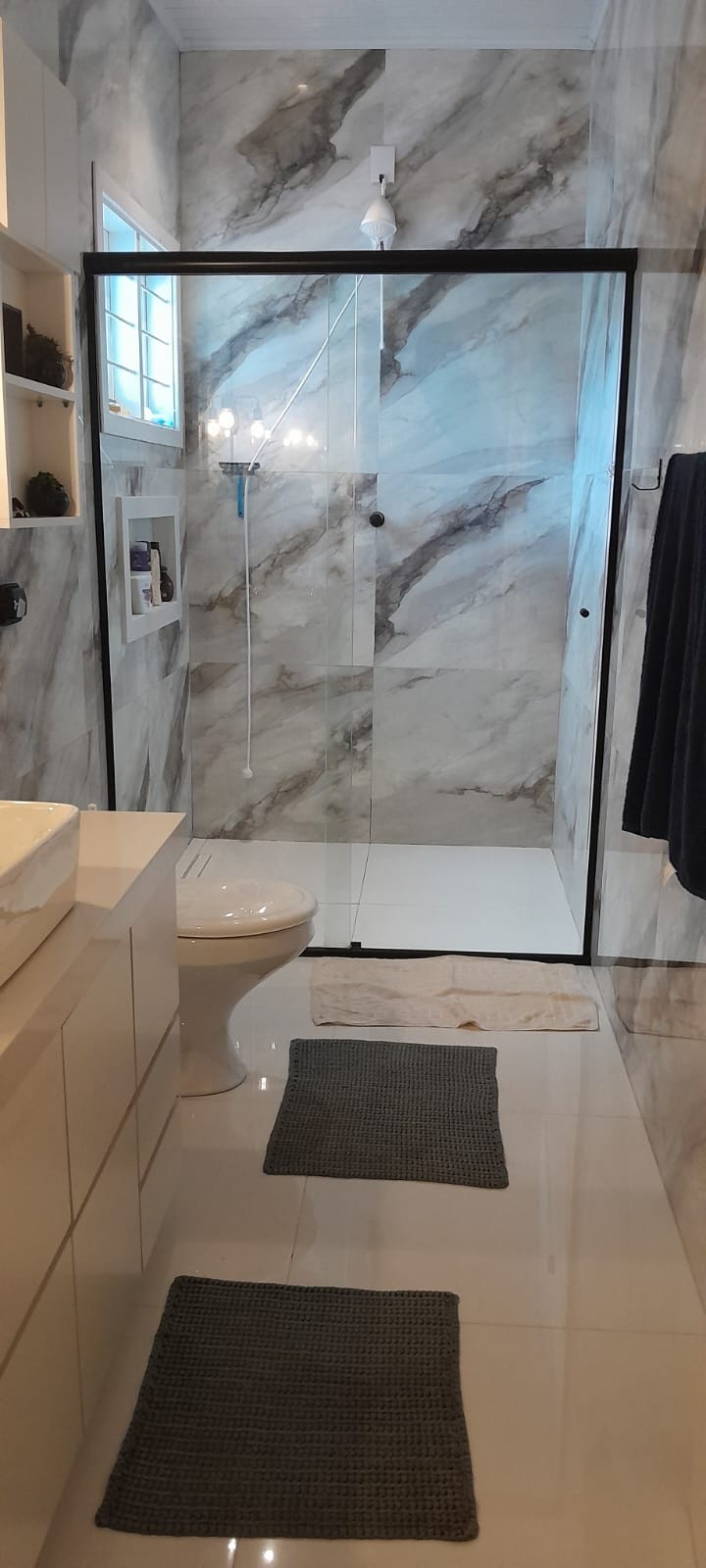 A sleek bathroom features marble-patterned walls and a glass-enclosed shower. A modern vanity with storage space is positioned beside a toilet. Neutral-toned bath mats are placed on the glossy floor, enhancing the clean and contemporary design.