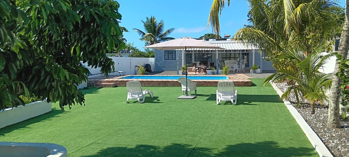 A well-maintained garden area features artificial grass, two sun loungers, and a large patio umbrella. The view is directed towards a swimming pool, with a seating area partially visible in the background. Lush palm trees enhance the tropical feel of the space.