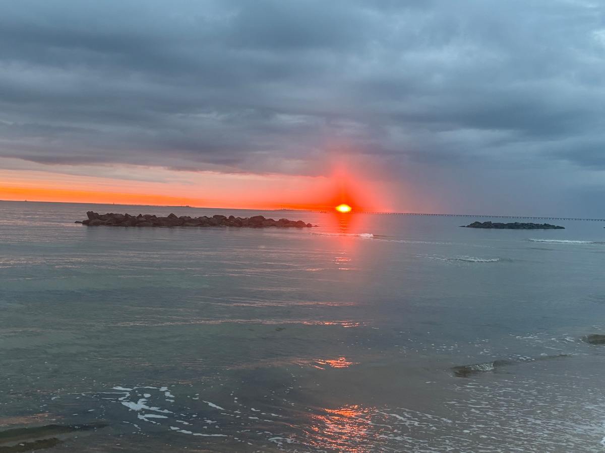 A serene beach scene captures the sun setting over the water, casting a warm glow across the horizon. Gentle waves create ripples along the sandy shore. Soft clouds in the sky add a touch of color to the tranquil evening atmosphere.