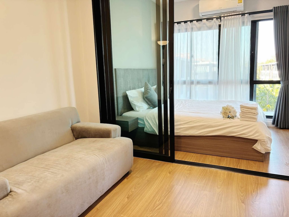 A cozy living area is highlighted by a light-colored sofa. A spacious bedroom area is visible through glass panels, featuring a neatly arranged bed with white linens. Natural light filters through sheer curtains, illuminating the wooden flooring.