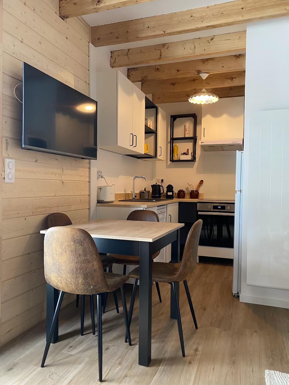 A modern kitchen area is presented, featuring wooden beams above and white cabinetry. A compact dining table is surrounded by four upholstered chairs. A television is mounted on the wall, and kitchen appliances and utensils are neatly arranged, showcasing a functional and inviting design.