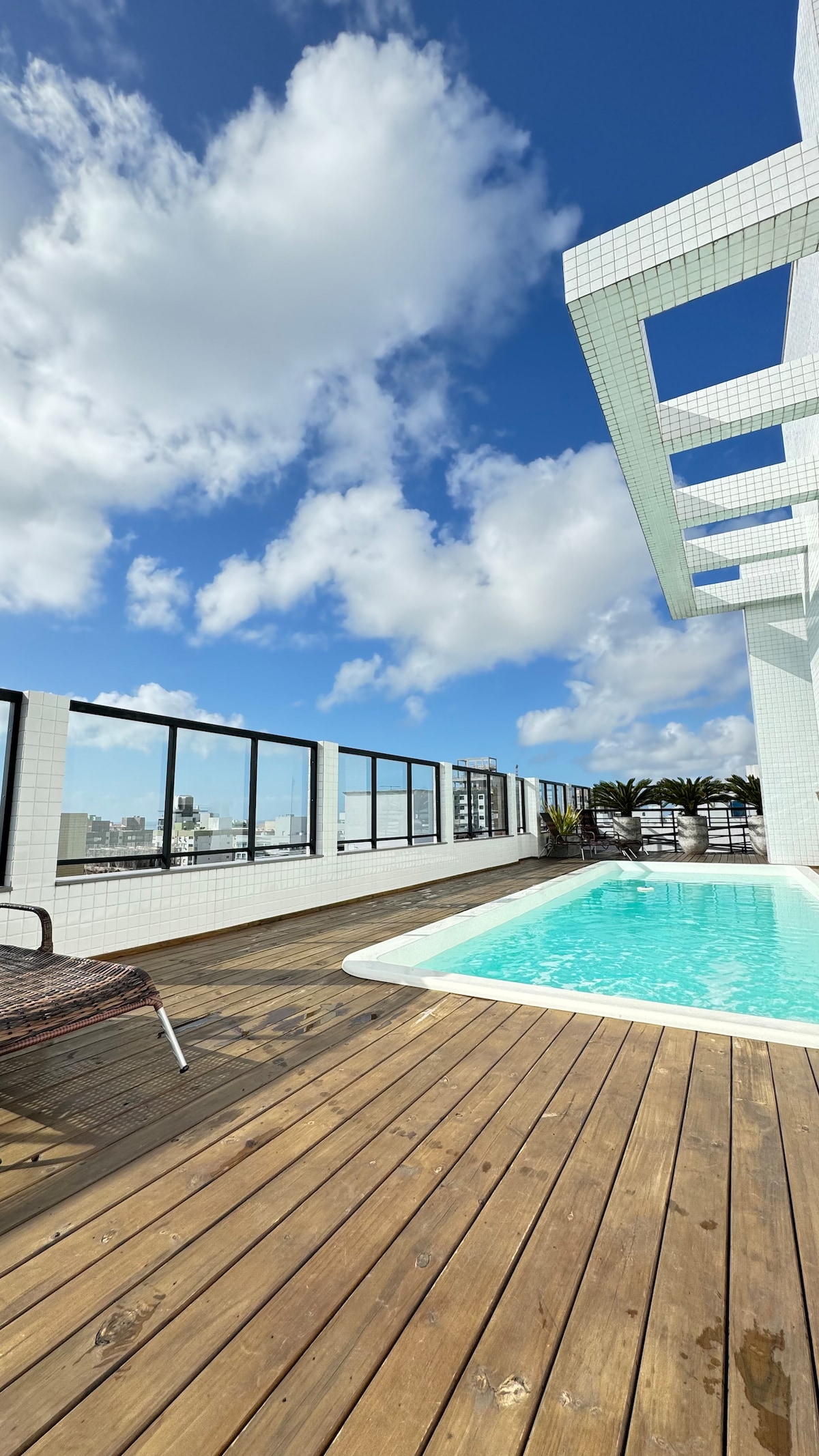The rooftop terrace features a refreshing pool surrounded by wooden decking. Glass railings provide unobstructed views of the skyline, while potted plants add greenery to the space under a bright blue sky dotted with clouds.