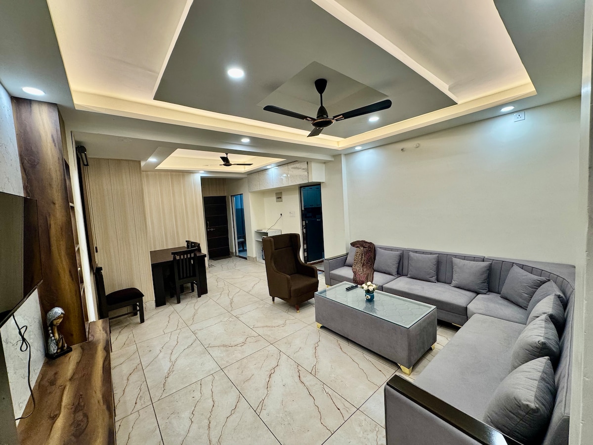 A spacious living area is furnished with a large sectional sofa and a central coffee table. Natural light fills the room from large windows, complementing the elegant ceiling design and subtle lighting. A dining table and chairs are positioned in the corner, enhancing the overall functionality.