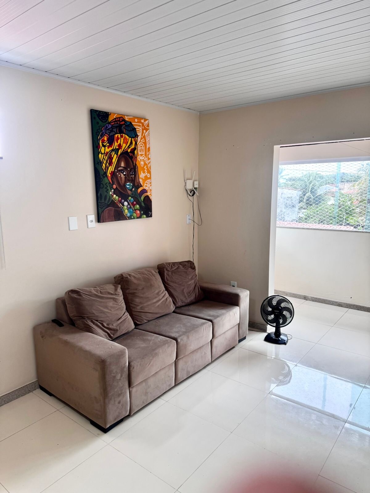 A cozy living area features a brown sofa positioned near a colorful artwork depicting a woman's face. Natural light enters through a nearby window, illuminating the polished tile floor. A small fan is located beside the sofa, adding comfort to the space.