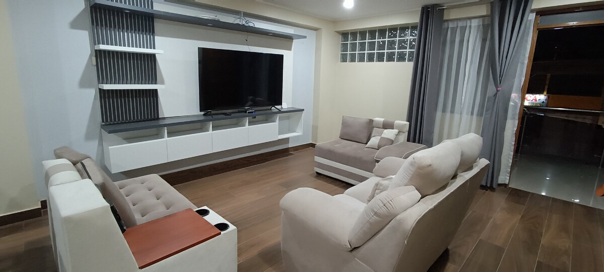 A modern living room is presented, featuring a large flat-screen television mounted on a wall unit. Two light-colored sofas are arranged around a central seating area, with a contrasting dark wooden floor. Natural light enters through a window, and sheer curtains enhance the space.