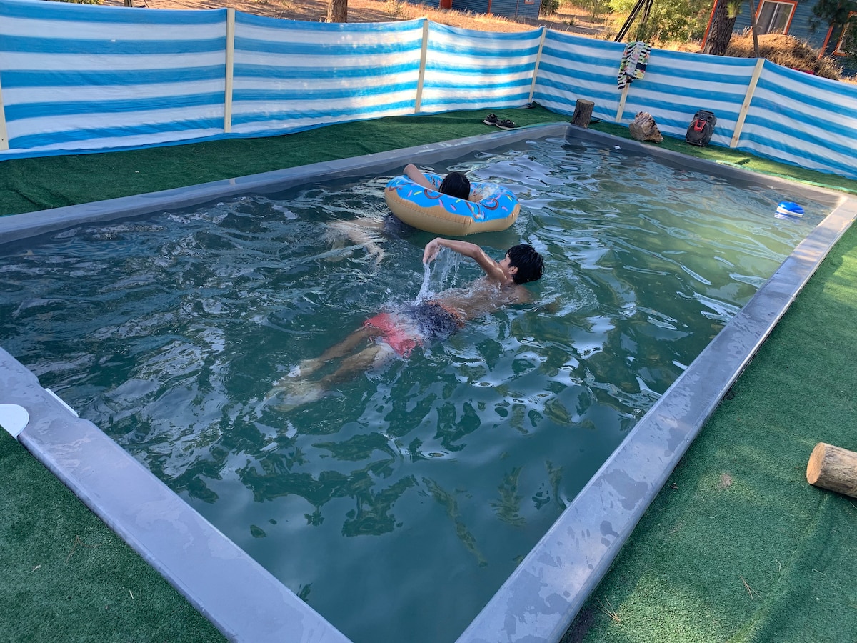 A rectangular pool is surrounded by a striped blue and white barrier. Two individuals are depicted in the water; one is swimming while the other floats on an inflatable ring. The pool is set on a grassy area, enhancing the outdoor experience.