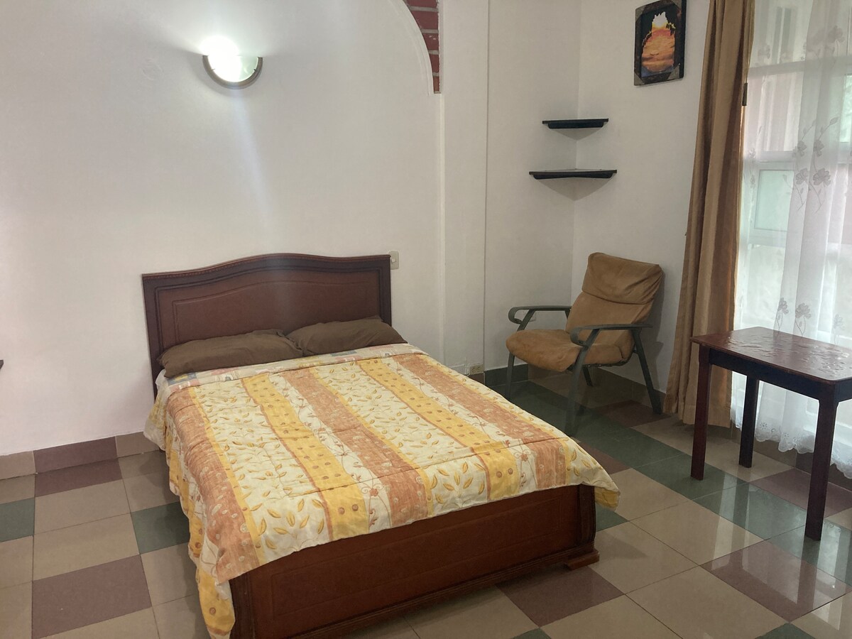 The sleeping area features a cozy double bed with a patterned yellow blanket, complemented by two brown pillows. A simple wooden nightstand and a chair are present, while a window adorned with sheer curtains allows for natural light to illuminate the space.