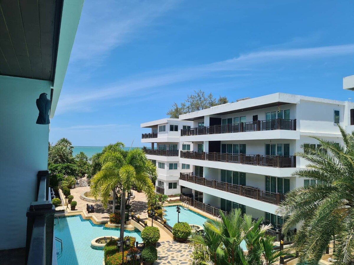 A view from a balcony showcases a serene outdoor area with well-maintained gardens and palm trees. The swimming pool is visible in the foreground, while modern buildings are lined along the coastline, offering a glimpse of the ocean and clear blue sky.