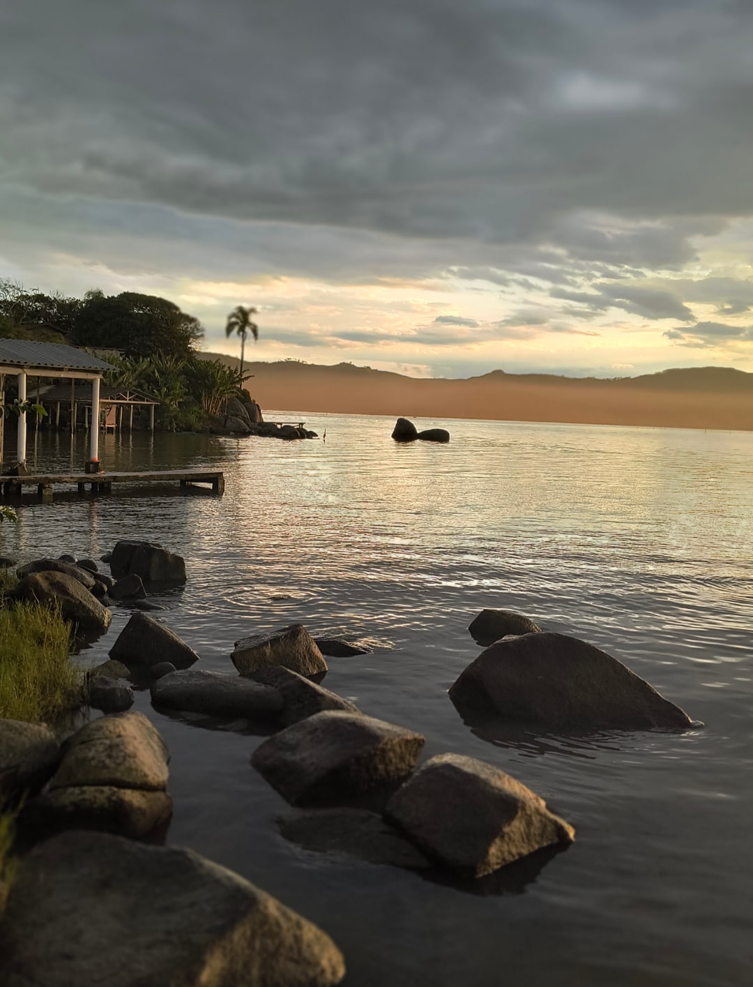 A serene waterfront scene is depicted, featuring smooth stones lining the shore. Calm waters reflect the soft colors of the sunset, while distant hills can be seen under a partly cloudy sky. A wooden structure is visible to the left, adding to the tranquil ambiance.