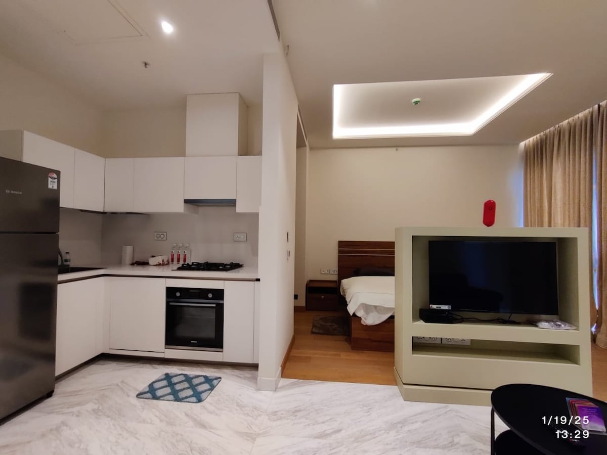 An open-plan living area features a modern kitchen with sleek cabinetry and essential appliances, including a refrigerator and stove. A spacious bed is positioned beyond the kitchen, while a flat-screen television is set in a minimalistic entertainment unit. Natural light flows through the window, complemented by soft curtains.