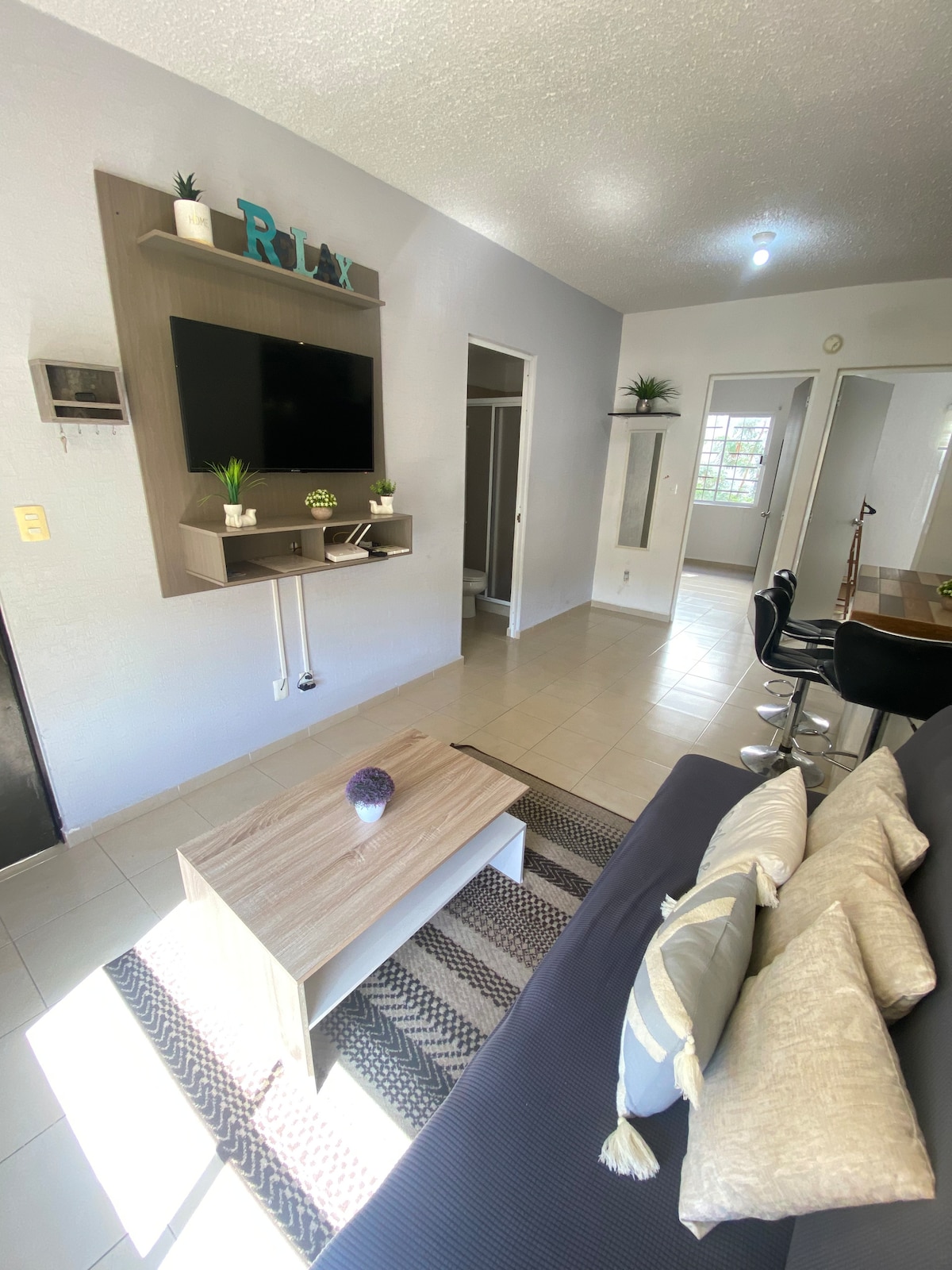 A living area is shown, featuring a neutral-colored sofa with decorative cushions. A wooden coffee table sits in front, while a wall-mounted television is positioned above a modern shelf. The space is illuminated by natural light coming from a nearby window.