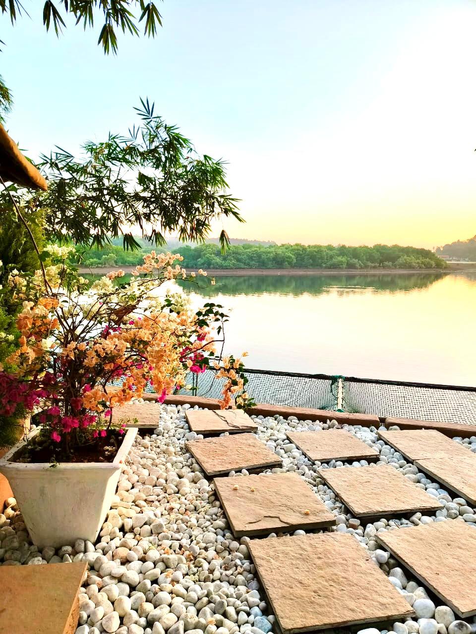 A serene outdoor space features a collection of stone tiles arranged over a bed of white pebbles. In the background, a tranquil lake reflects soft colors of sunrise, framed by lush trees. Potted flowering plants add a touch of color to the setting.